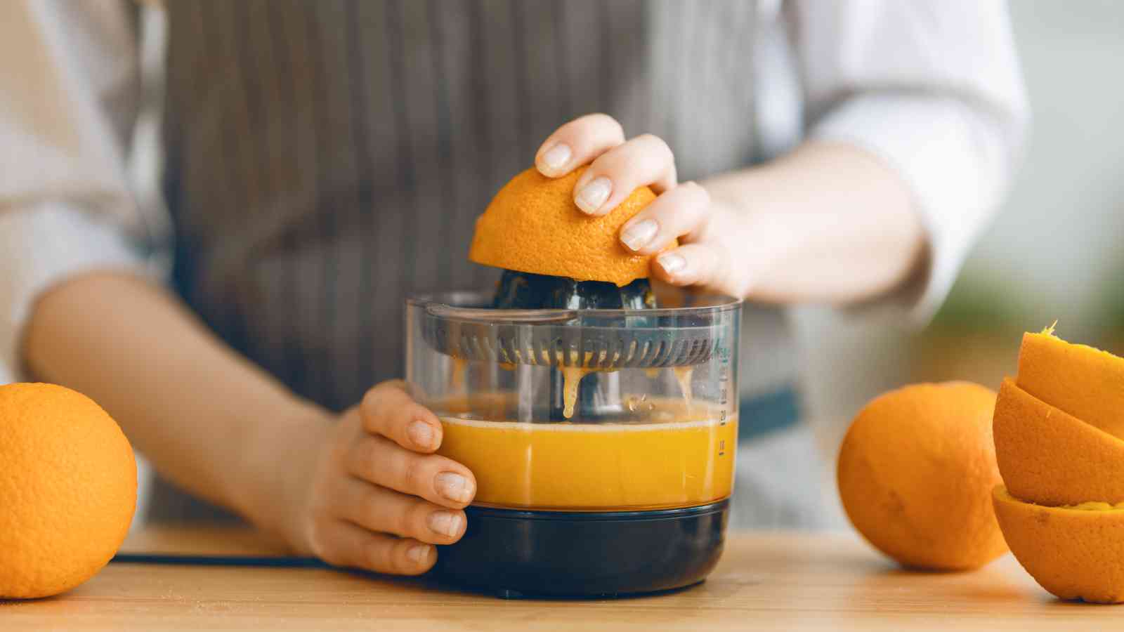 A person uses a manual juicer to squeeze juice from an orange, with several whole and halved oranges on a wooden surface nearby.