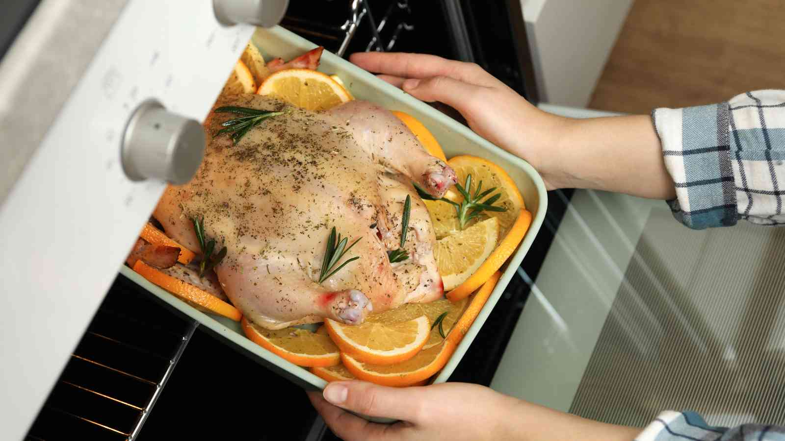 A person places a raw, seasoned whole chicken on a bed of orange slices into an oven using a rectangular baking dish.