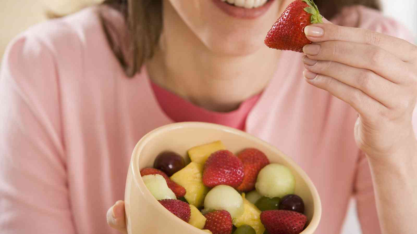 A person holding a bowl of mixed fruit, including strawberries, grapes, pineapple, and melon, and lifting a strawberry to their mouth.
