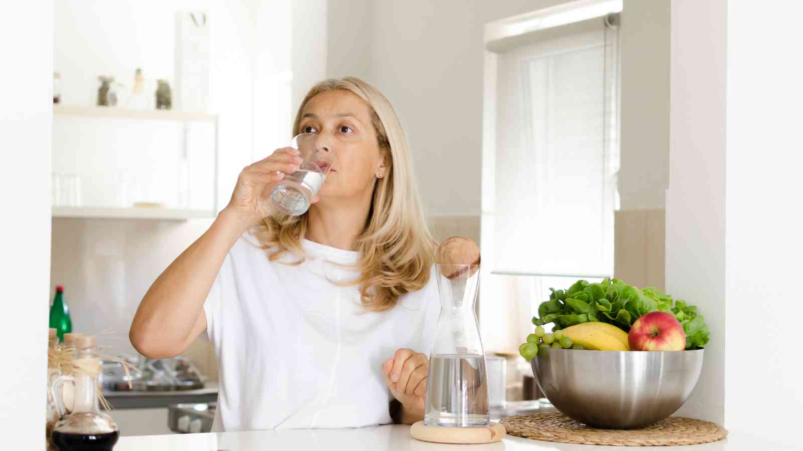 A woman stands in a kitchen, drinking water from a glass. A carafe of water and a bowl of fresh fruits and vegetables are on the counter in front of her.