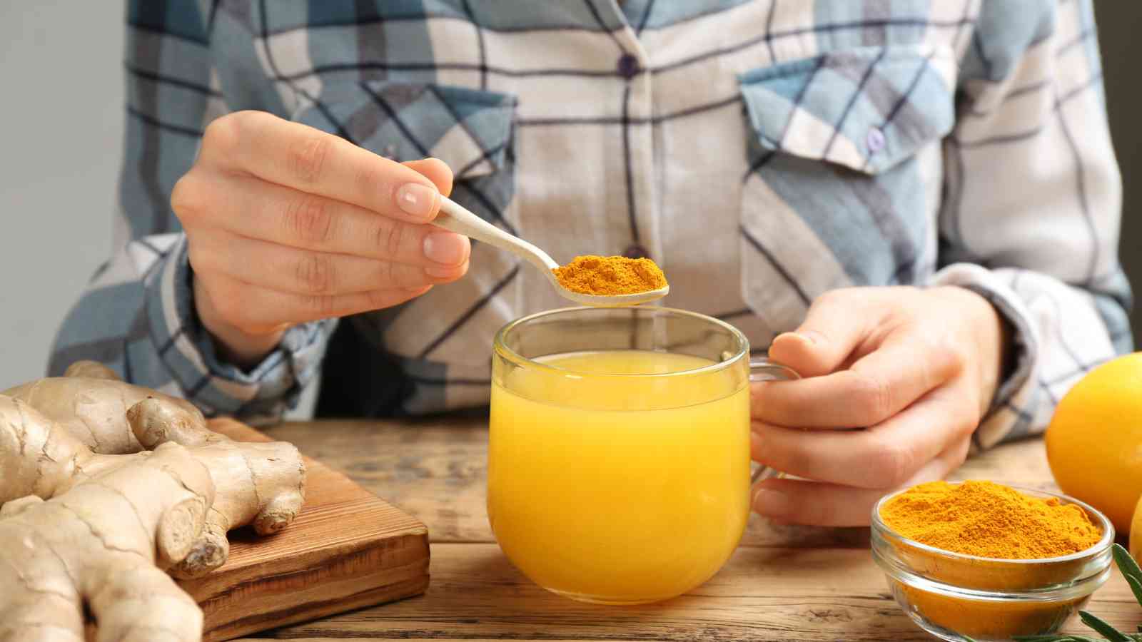 Person adds a spoonful of turmeric powder to a cup of yellow liquid, with ginger, a bowl of turmeric, and a lemon on a wooden table.