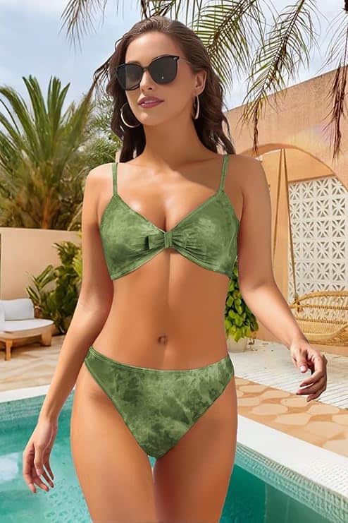 A woman wearing sunglasses and a green bikini stands by a pool, with palm trees and outdoor furniture in the background on a sunny day.
