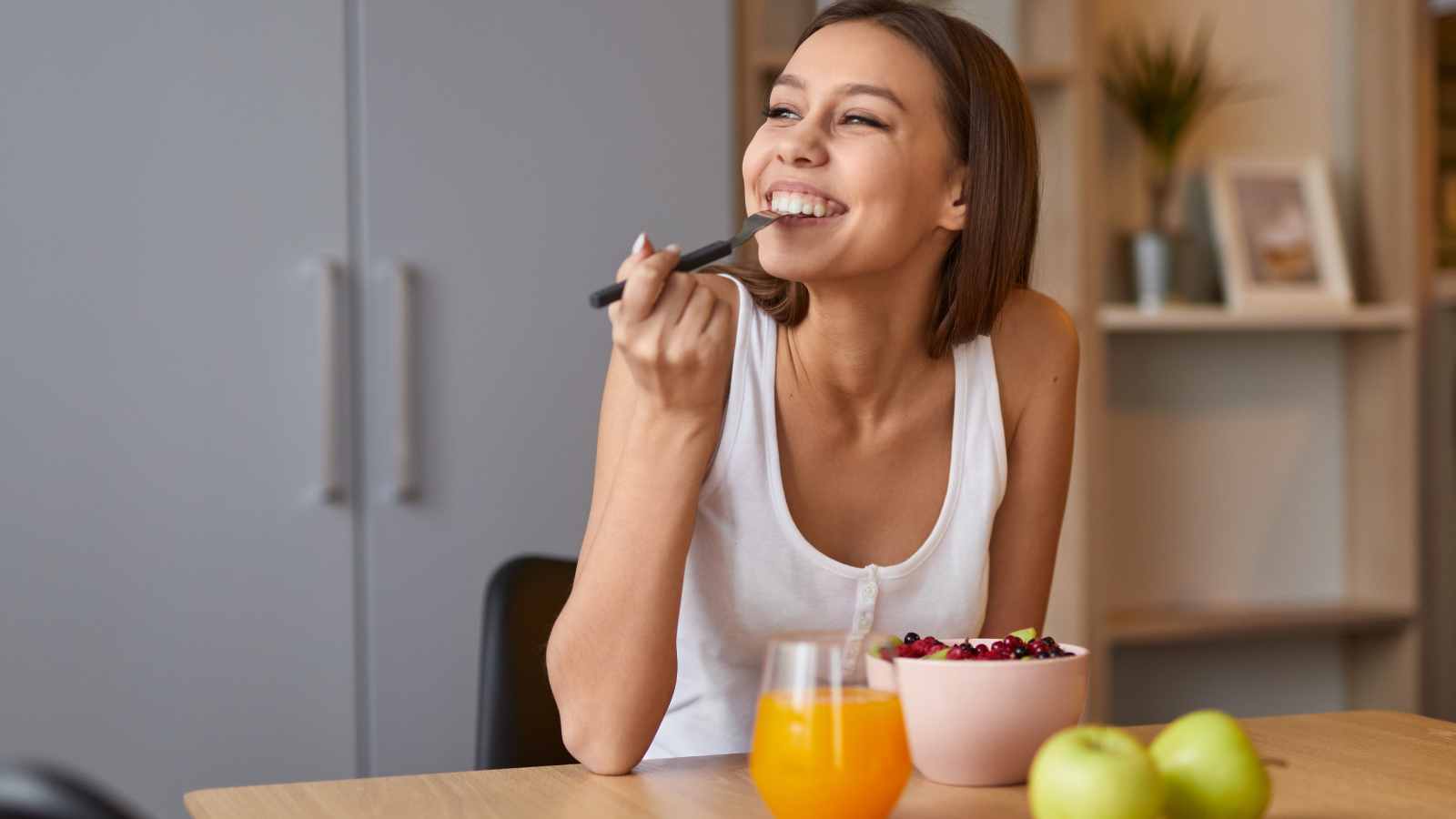 Woman in a white tank top smiles while eating from a bowl of fruit at a table with a glass of orange juice and green apples.