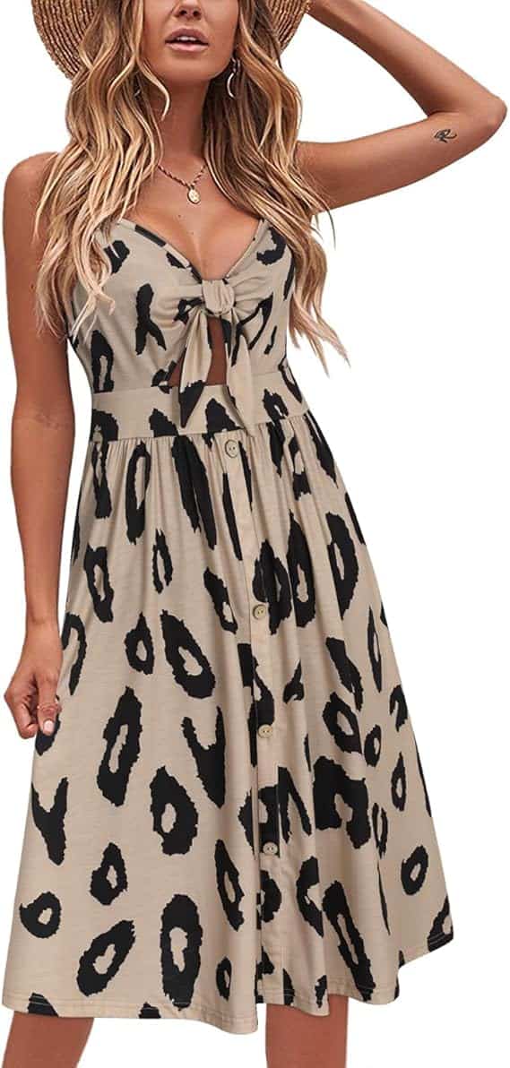 Woman wearing a sleeveless beige dress with a black leopard print pattern, front tie detail, button accents, and a flared skirt. She has long wavy hair and is touching a wide-brimmed hat.