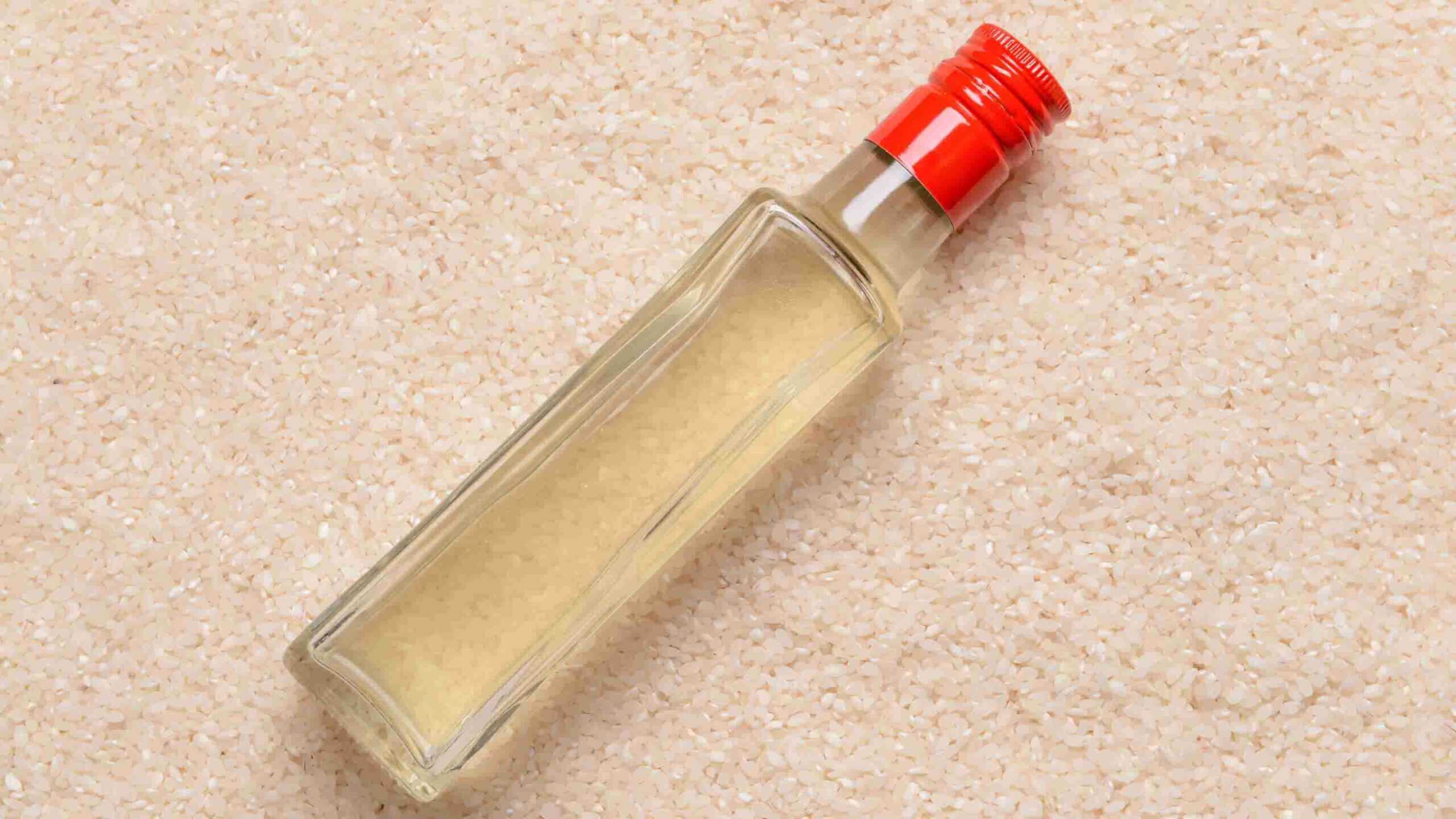 A clear glass bottle with a red cap filled with a pale liquid, lying on a surface covered with uncooked white rice grains.