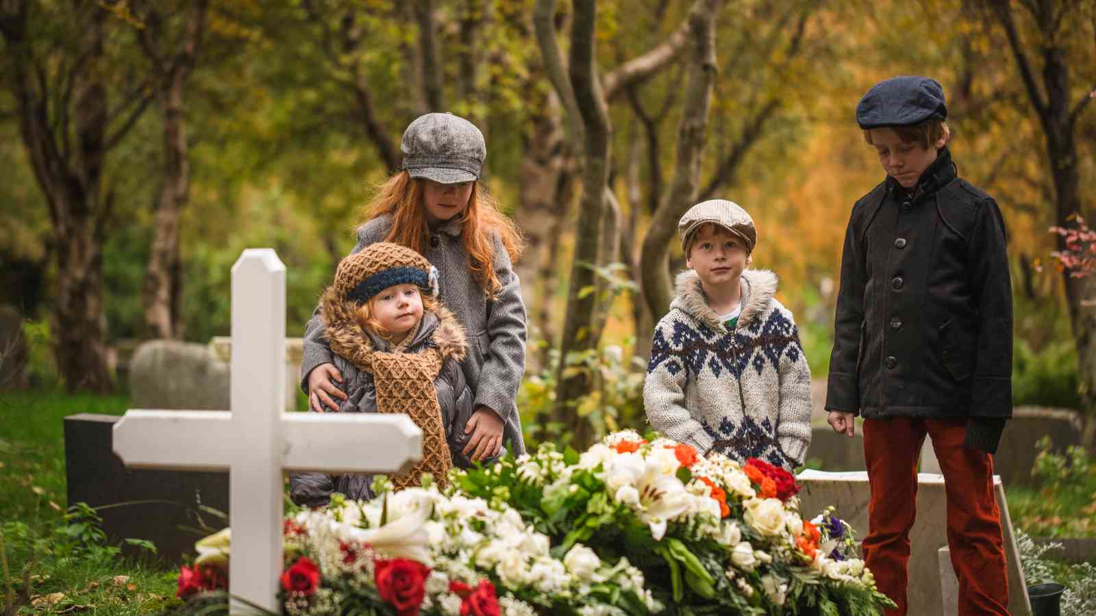 Four children stand by a white cross grave marker with flowers in a cemetery, surrounded by trees in autumn.