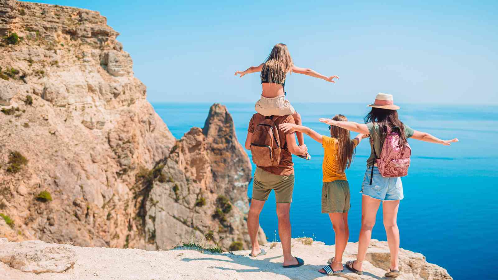 A family of four stands on a cliff edge overlooking the sea, with three members extending their arms outward and one child sitting on an adult’s shoulders.