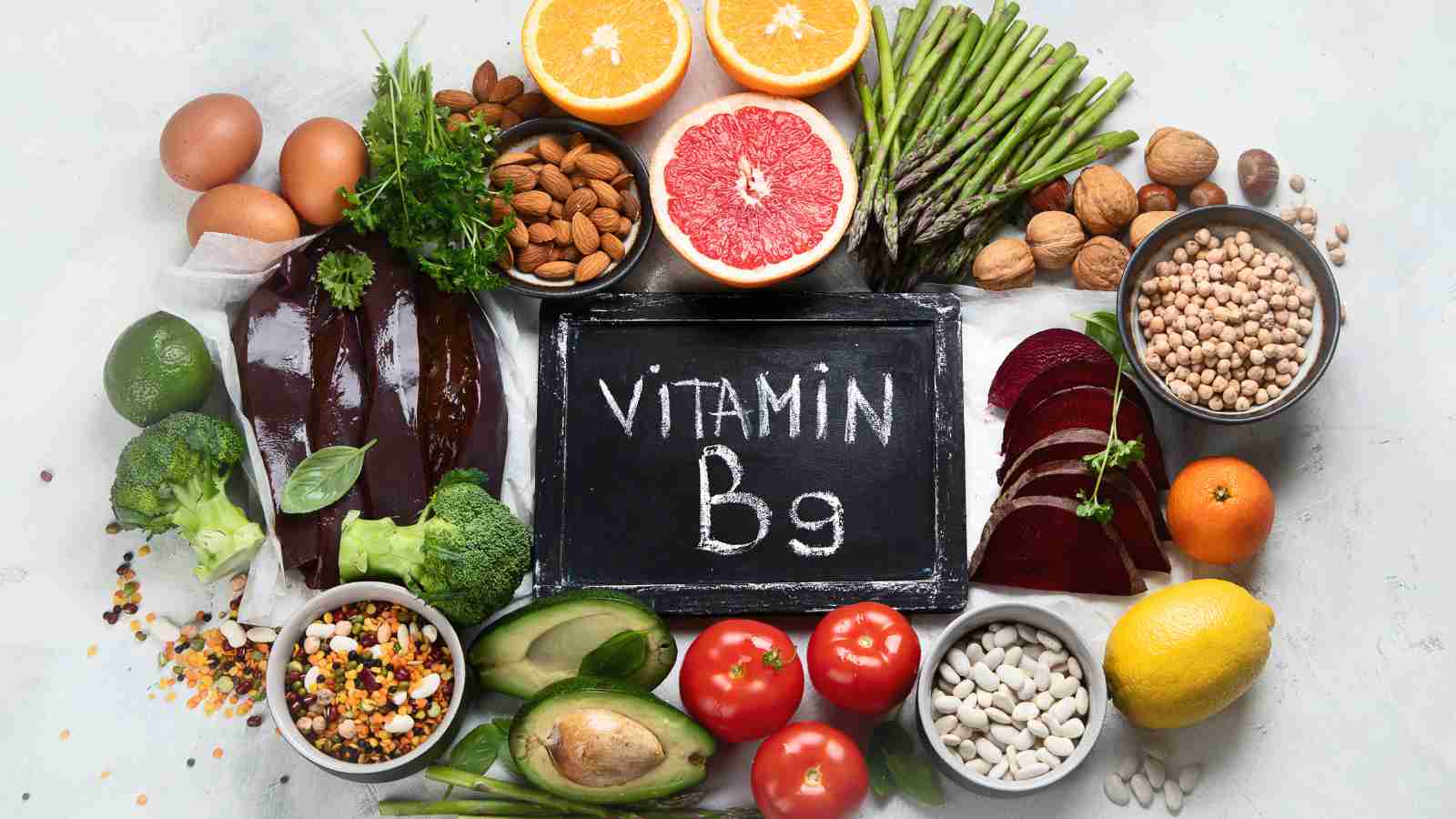 A variety of vitamin B9-rich foods, including fruits, vegetables, legumes, nuts, and eggs, are arranged around a small chalkboard with the text "Vitamin B9".