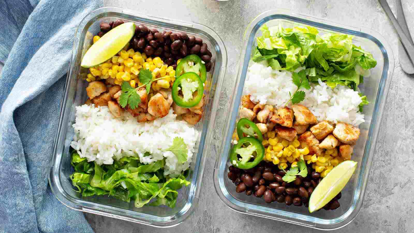 Two glass meal prep containers filled with white rice, black beans, corn, diced chicken, lettuce, lime wedges, jalapeño slices, and cilantro on a light surface.