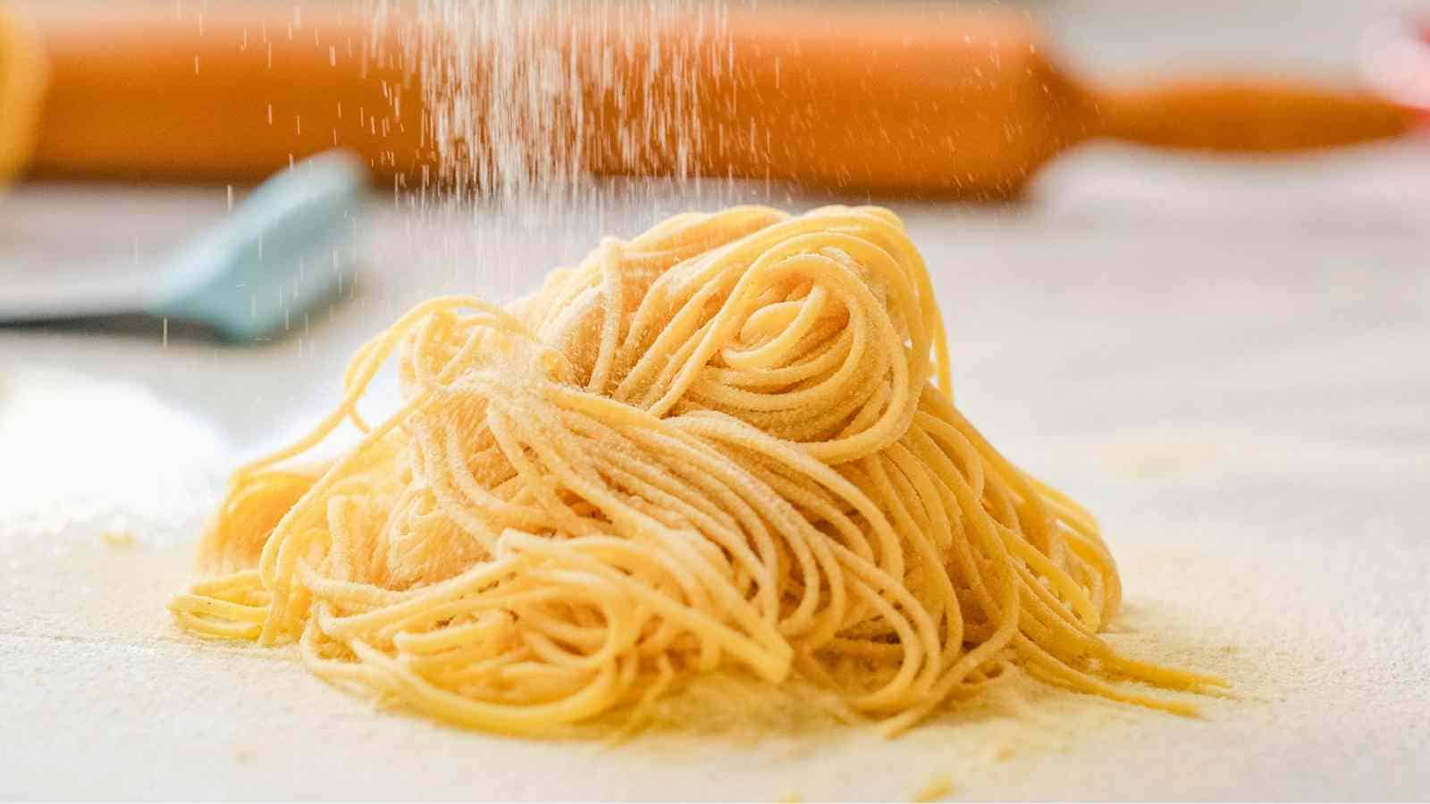 A pile of raw spaghetti noodles on a floured surface with flour being sprinkled on top; a rolling pin and spatula are blurred in the background.