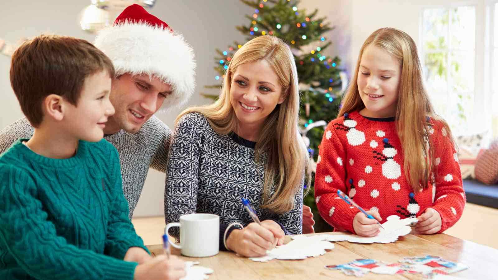 A family of four sits at a table making holiday crafts, with a Christmas tree and festive decorations in the background.