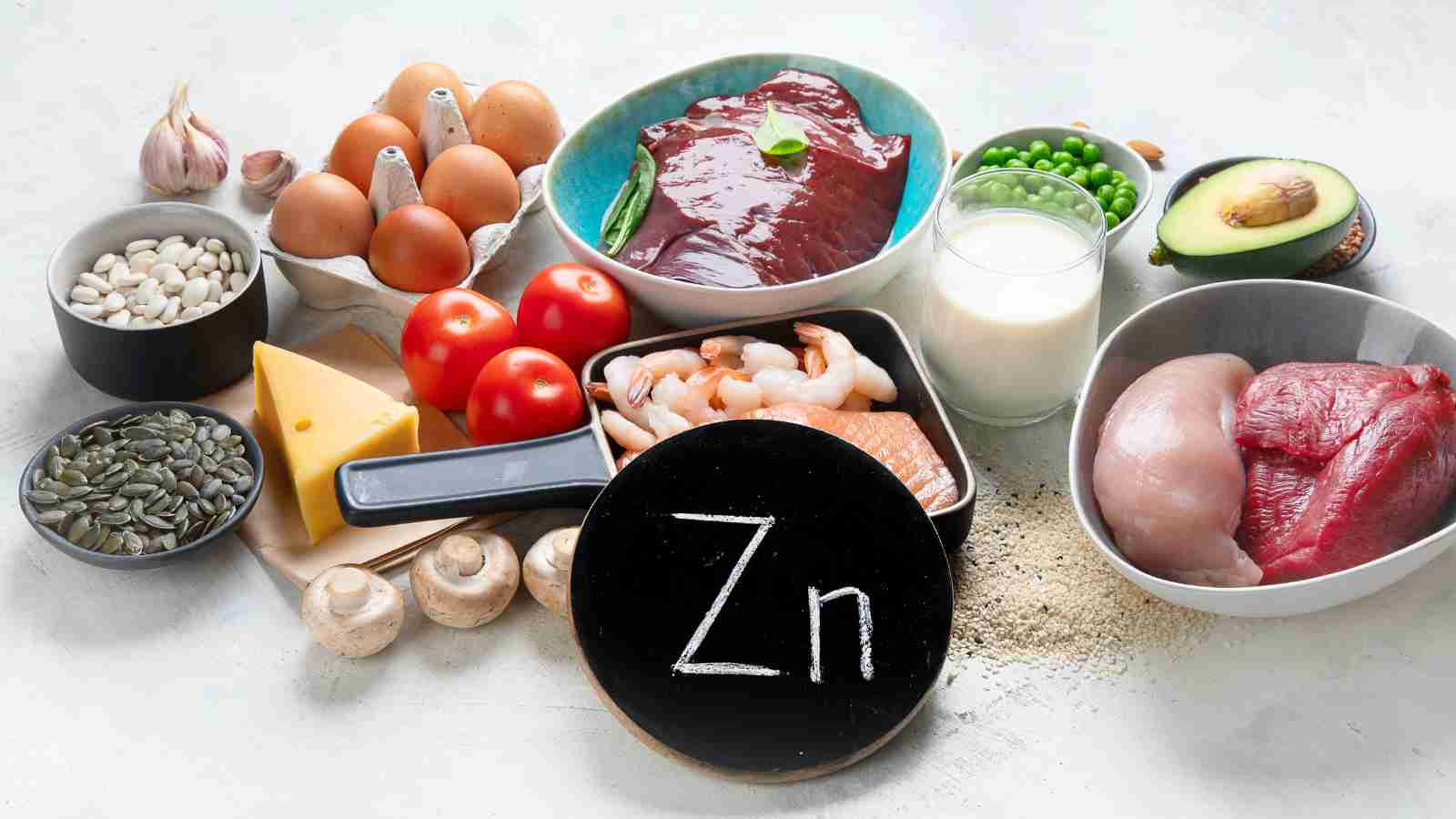 A variety of zinc-rich foods, including beef, chicken, eggs, nuts, seeds, and dairy, are arranged around a small chalkboard with the chemical symbol for zinc.