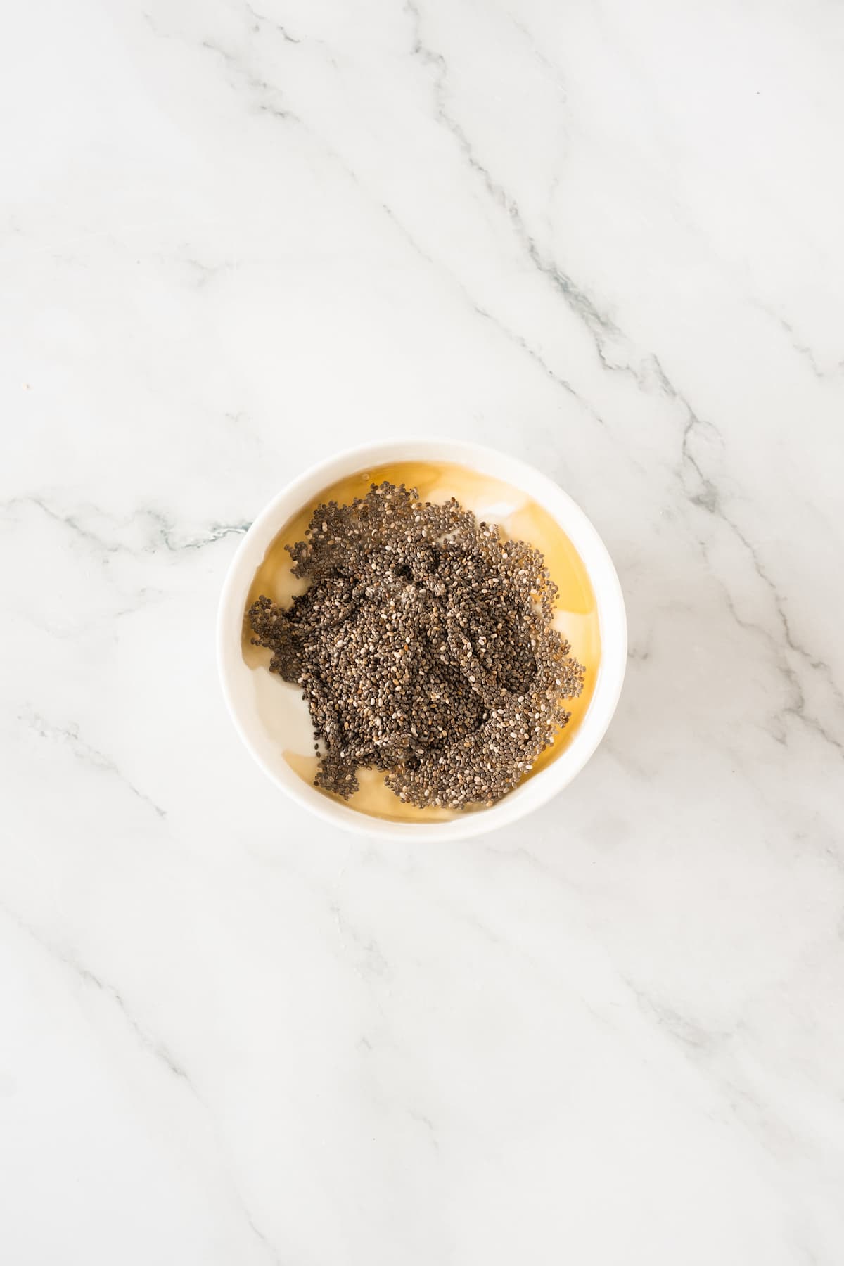 A white bowl containing agave syrup and chia seeds sits on a white marble surface.