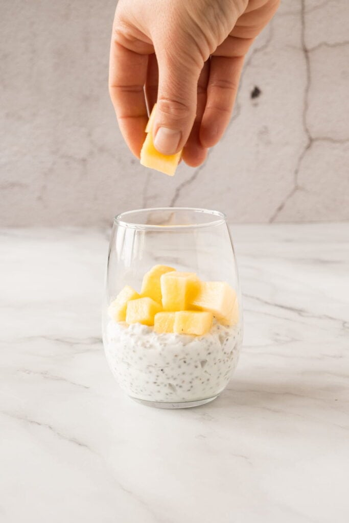 A hand adds a piece of pineapple to a glass of chia pudding topped with pineapple chunks on a marble surface.