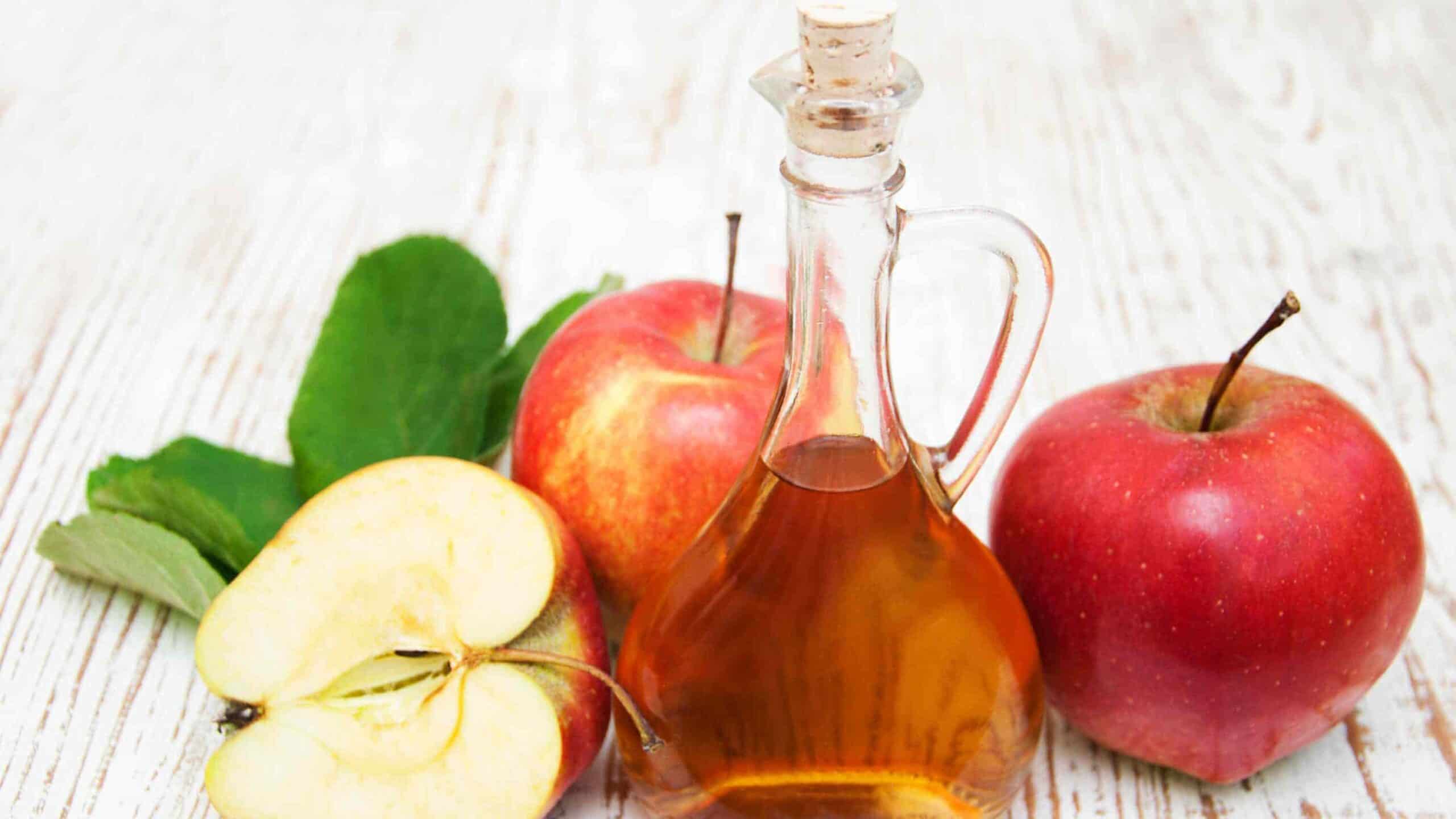 A glass bottle of apple cider vinegar sits on a light wooden surface next to two whole apples, apple slices, and green leaves.