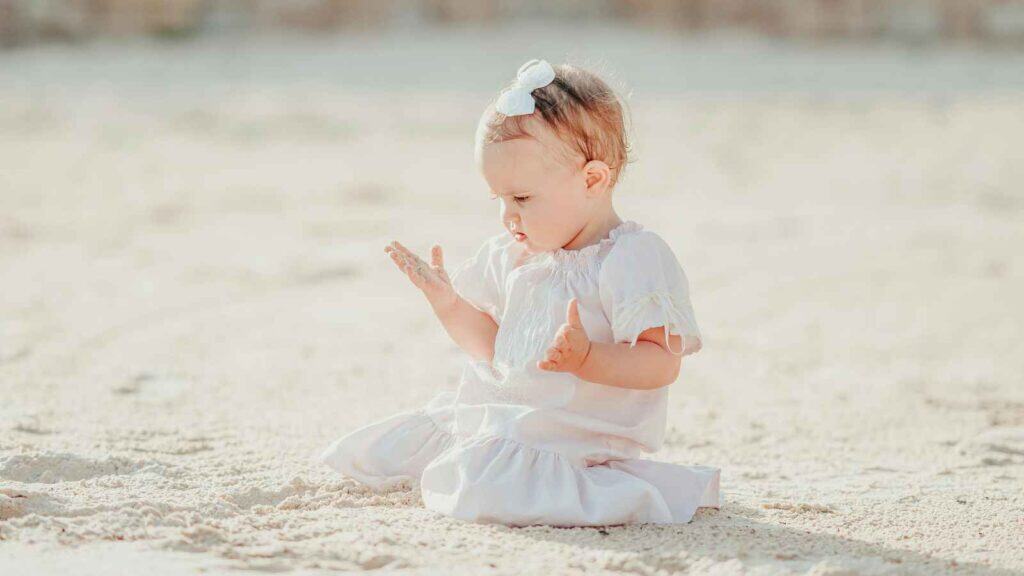 A baby wearing a white dress and headband sits on sandy ground, looking at their hands with a curious expression.