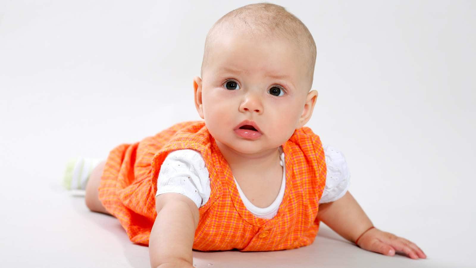 A baby wearing an orange dress and white shirt lies on their stomach on a light-colored surface, looking slightly upward.