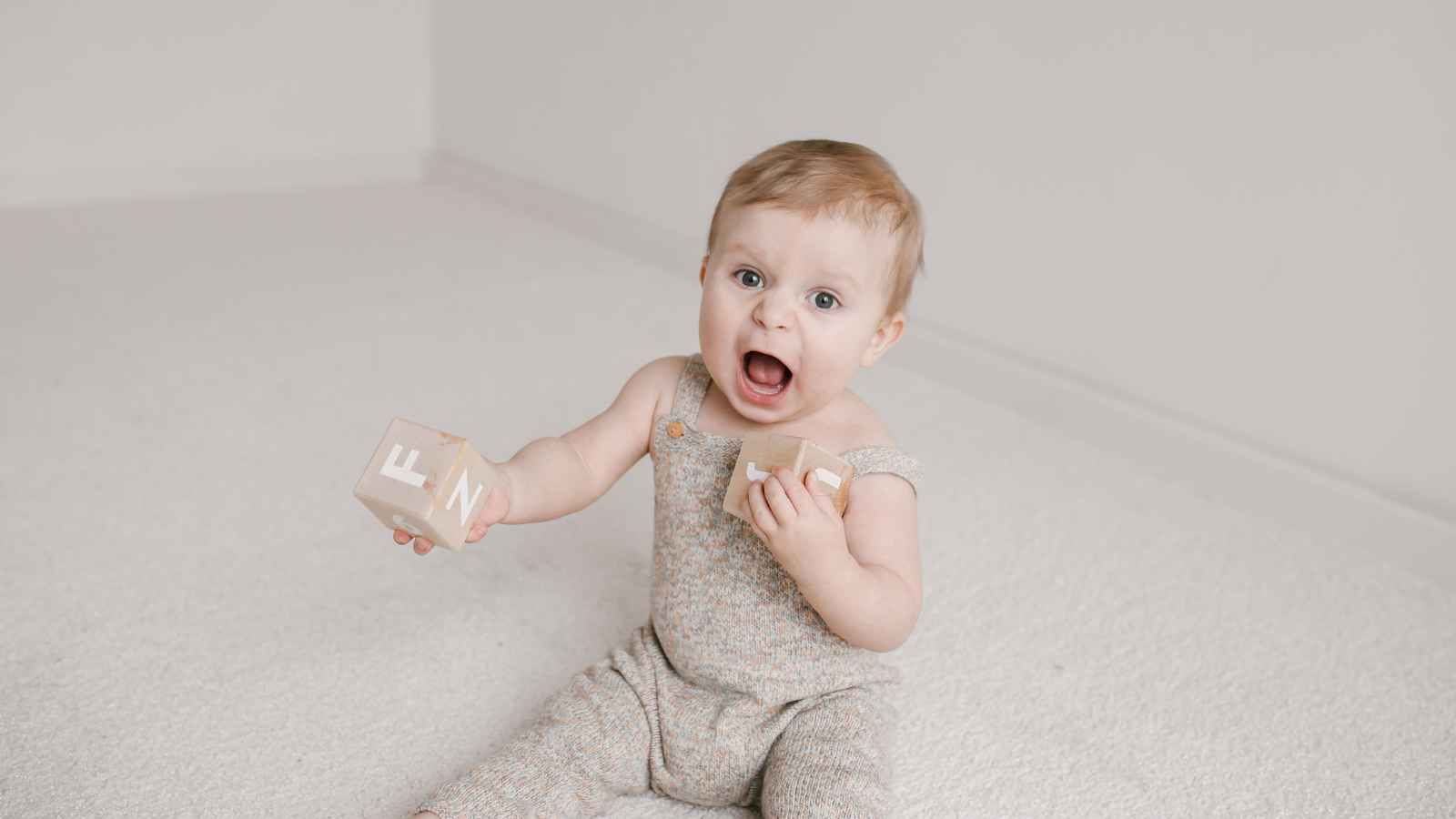 A baby sits on a light carpeted floor, wearing a knit romper, holding two wooden blocks, and looking up with an open mouth expression.