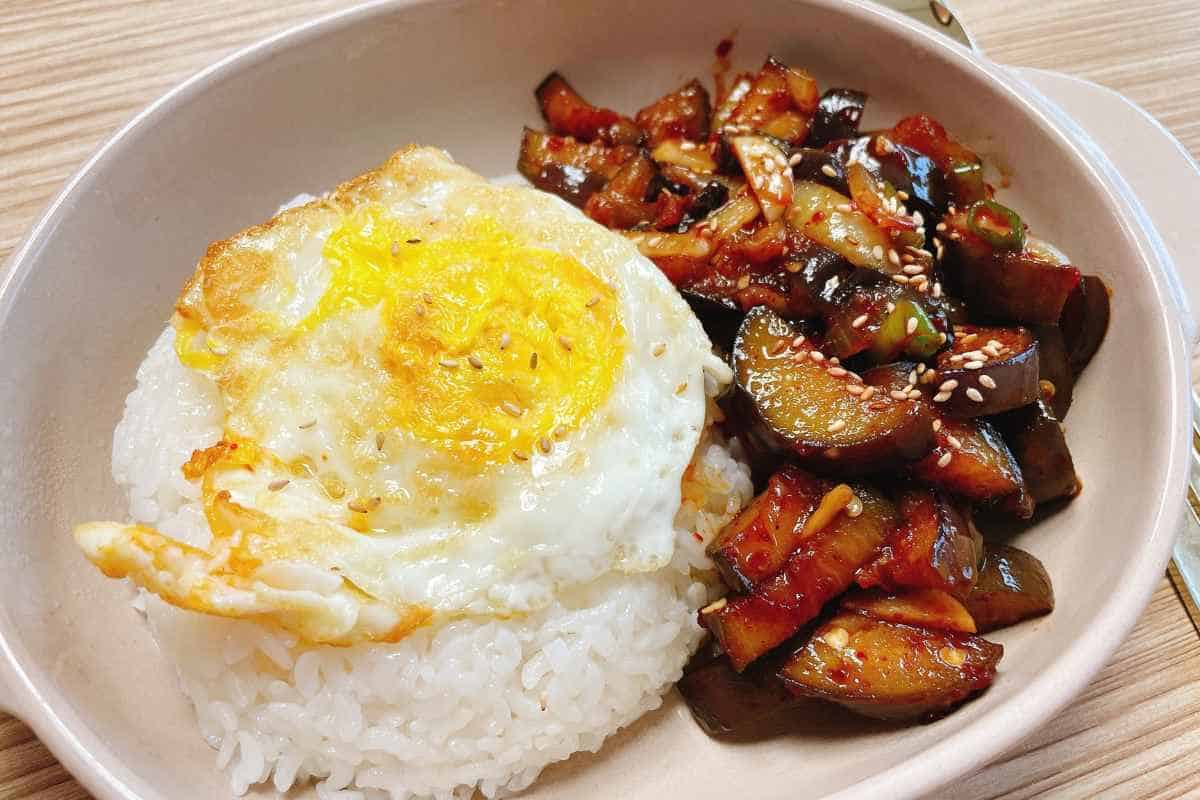 A bowl with white rice topped with a fried egg, served alongside stir-fried eggplant garnished with sesame seeds.