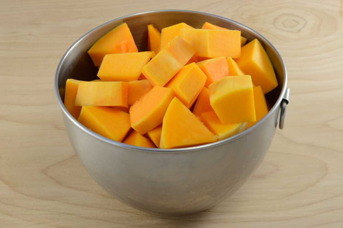A metal bowl filled with chunks of raw butternut squash sits on a light wooden surface.