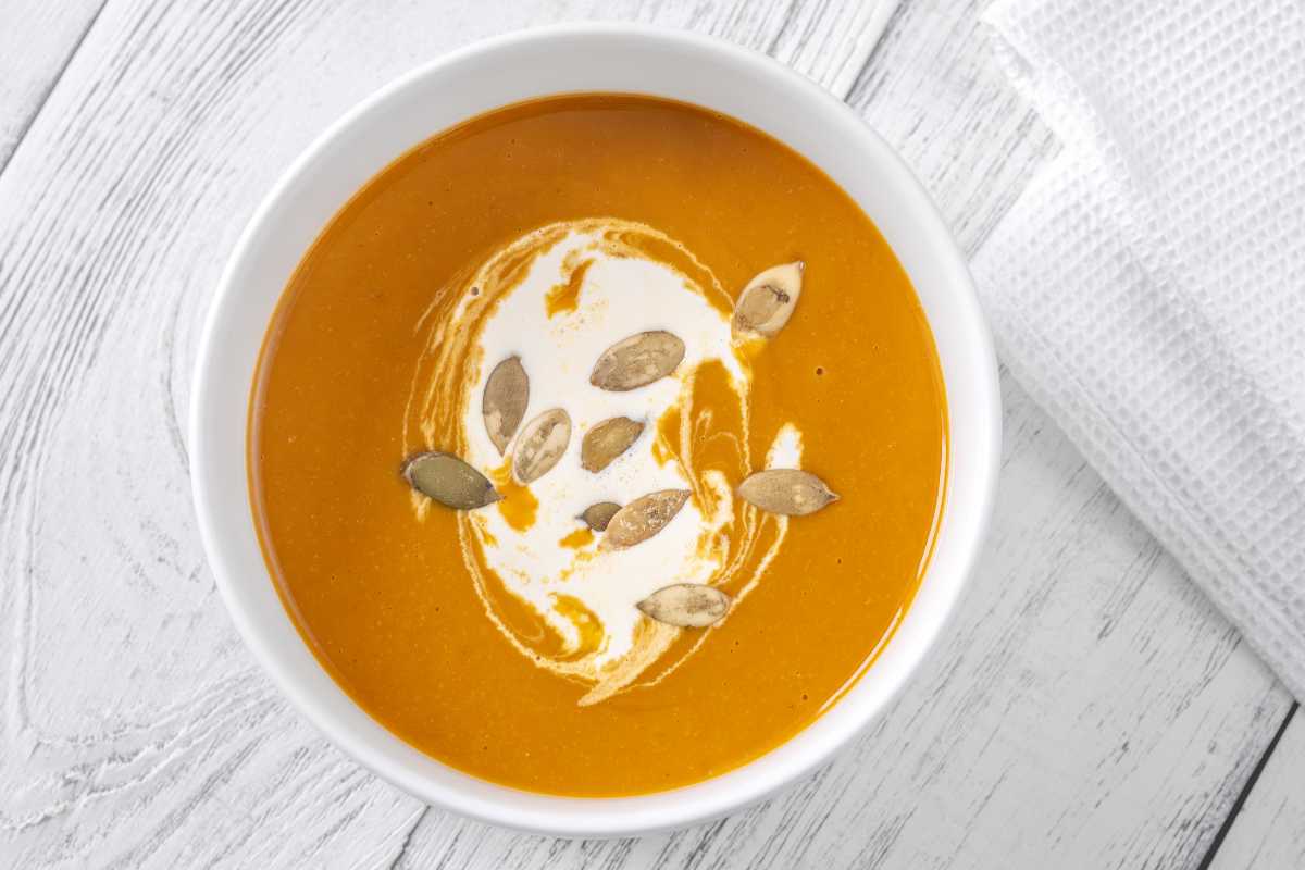 A white bowl of orange-colored soup garnished with a swirl of cream and pumpkin seeds, placed on a white wooden surface next to a white cloth.