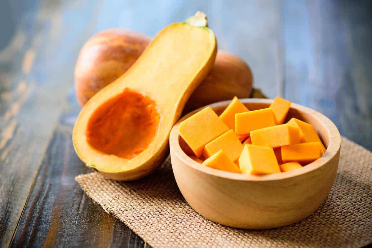 A halved butternut squash with seeds removed sits next to a wooden bowl filled with cubed squash on a burlap mat.