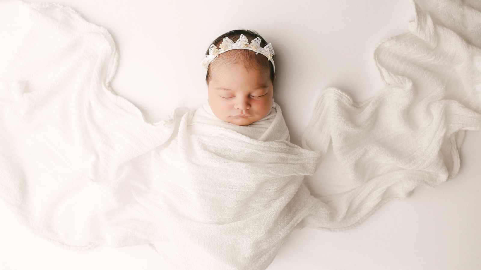 A baby wrapped in a white blanket wearing a floral headband.