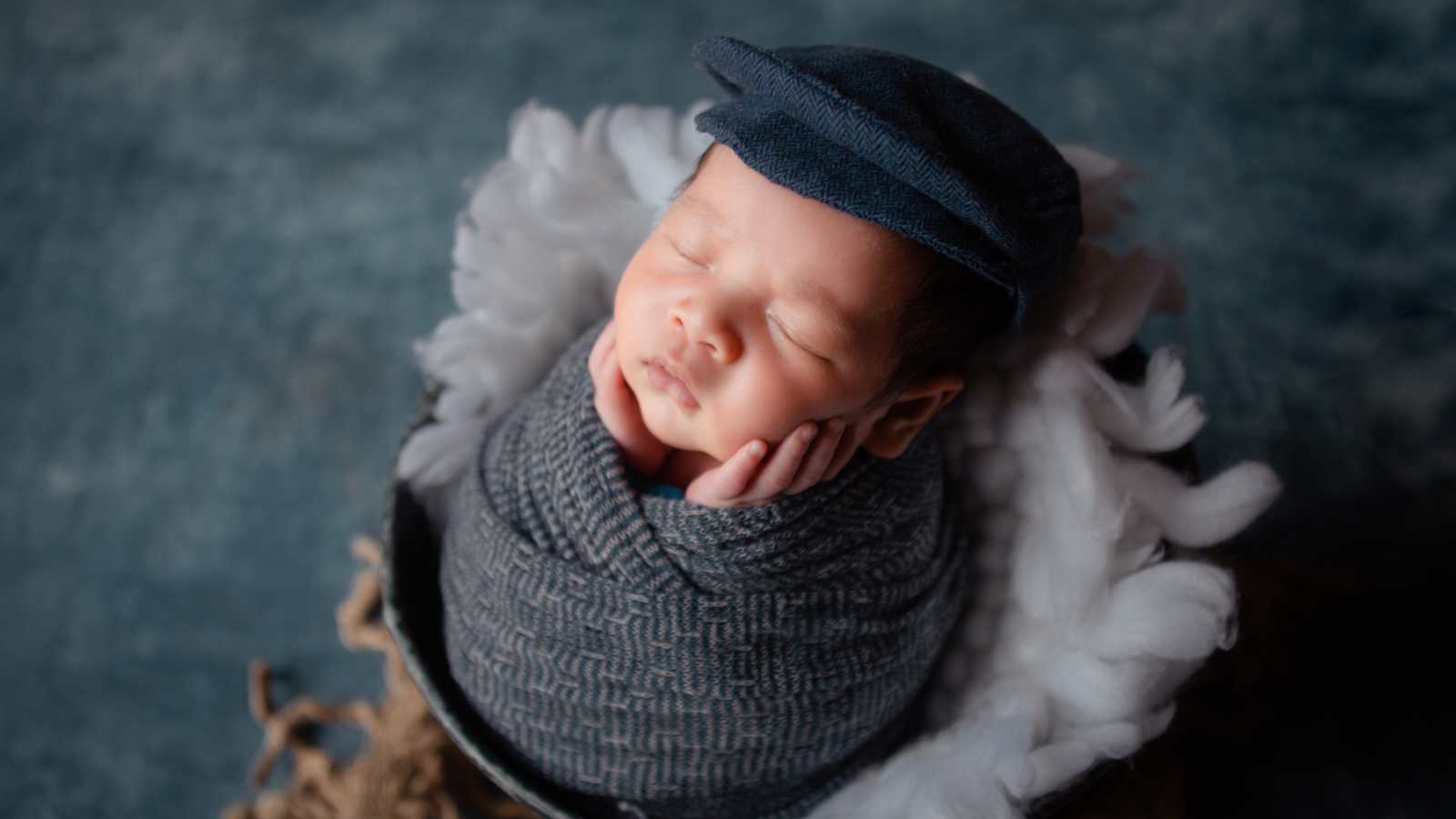 Newborn baby wrapped in a gray blanket and wearing a cap, sleeping peacefully with hands on cheeks, surrounded by soft white padding.