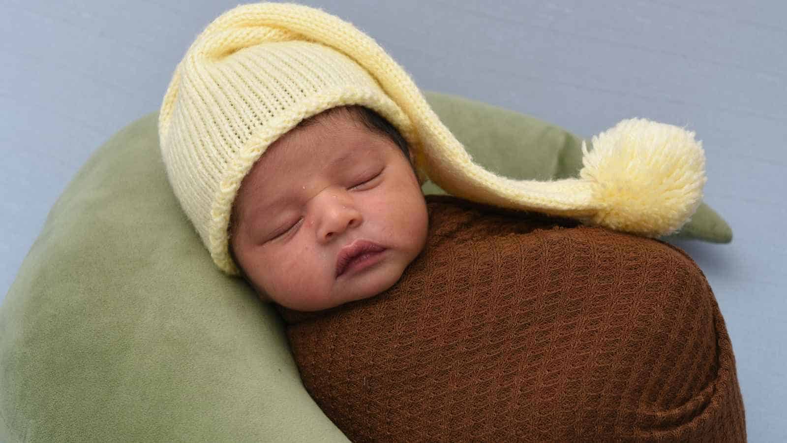 Newborn baby sleeping, wrapped in a brown blanket, wearing a yellow knit hat with a pom-pom, resting on a green cushion.