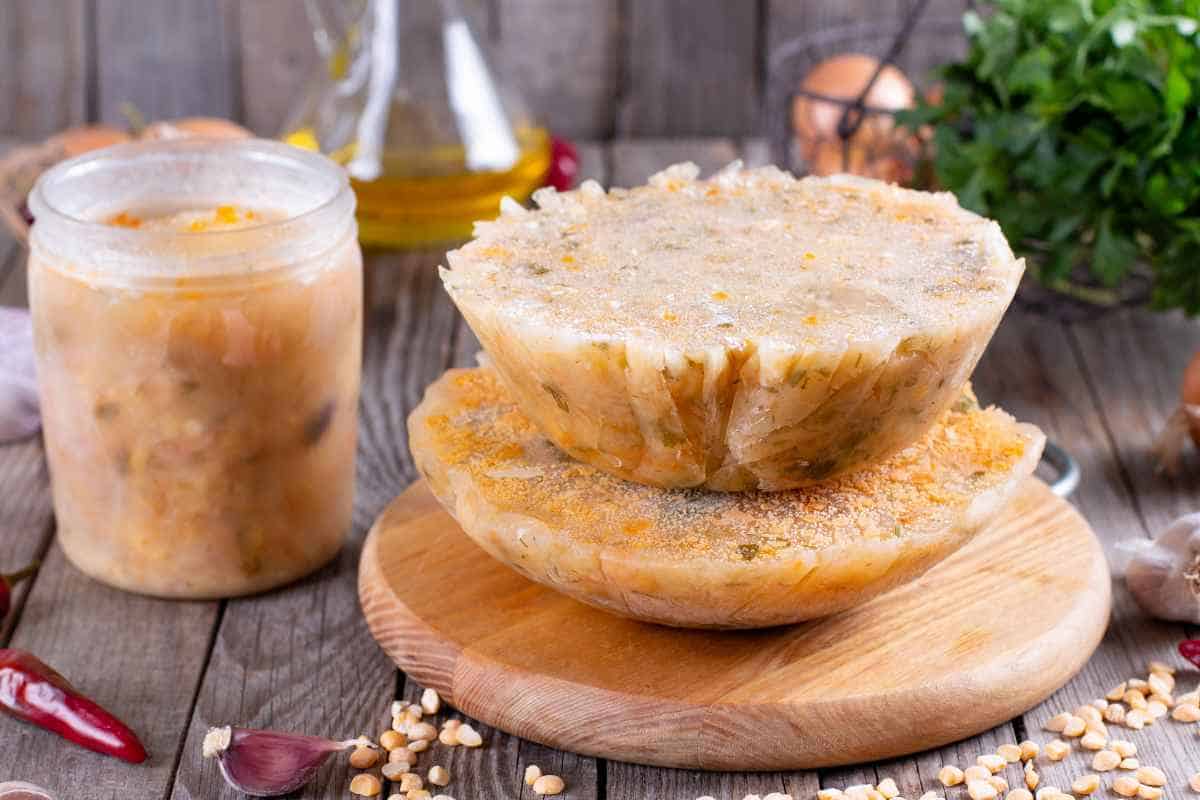 Two stacked bowls of jellied meat, known as aspic, rest on a wooden board. A jar of aspic and various ingredients are visible in the background.
