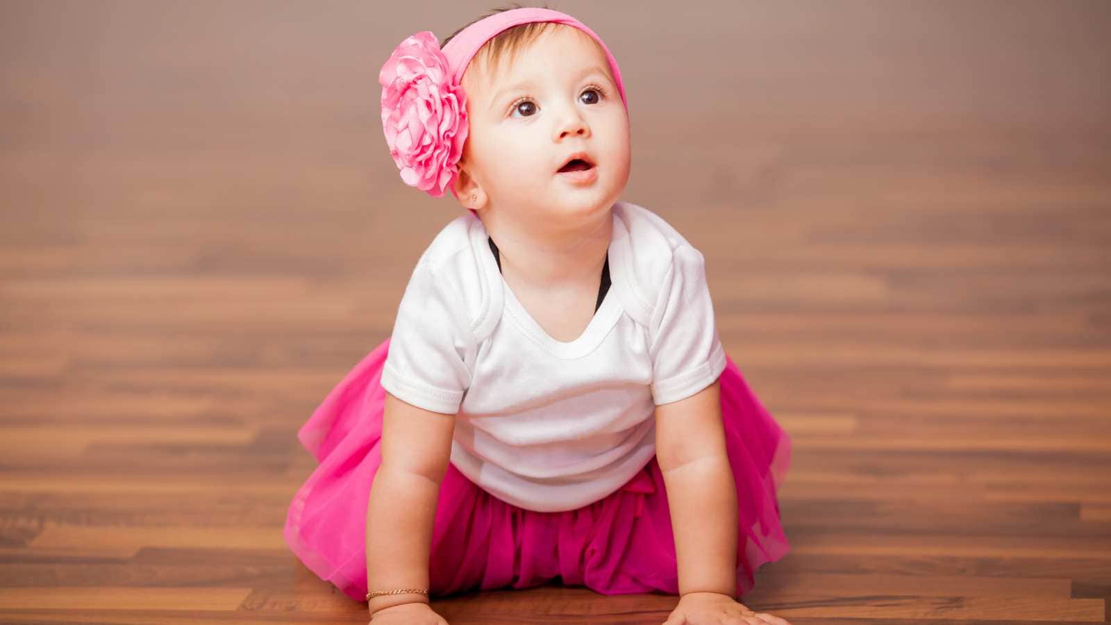 A baby wearing a white shirt, pink tutu, and pink headband with a large flower crawls on a wooden floor, looking upward.