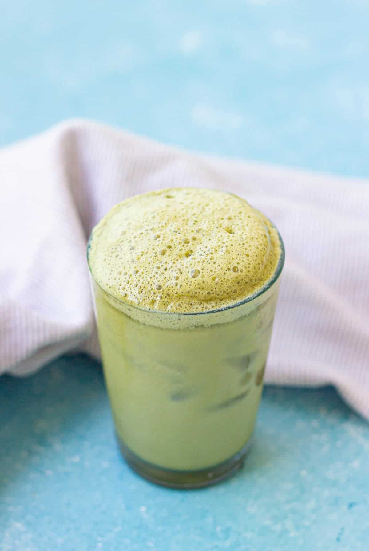 A glass of iced matcha latte with a frothy green top sits on a blue surface, next to a folded light-colored cloth.