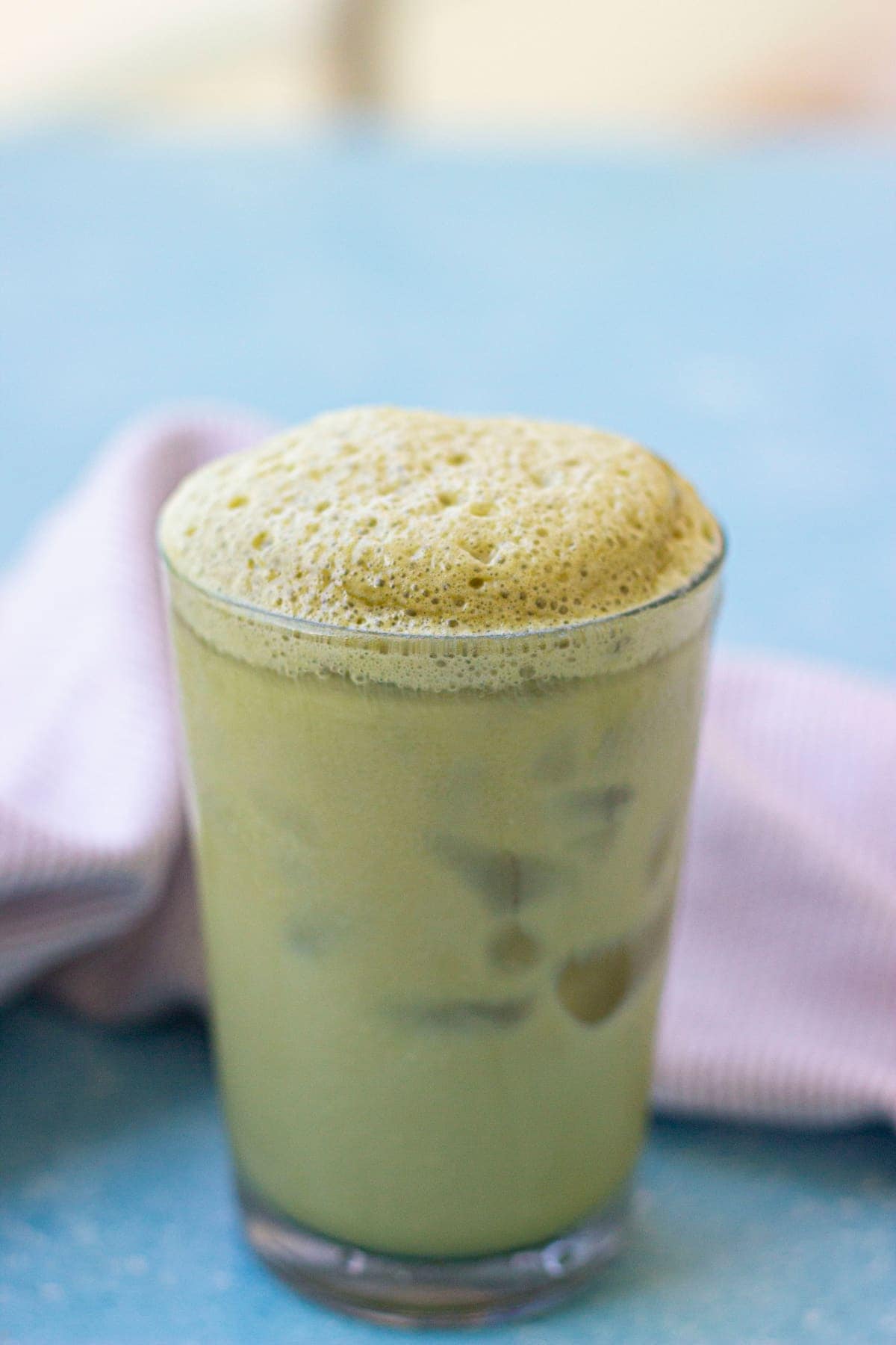 A glass of iced matcha latte with a thick layer of frothy foam on top, placed on a blue surface next to a folded light purple cloth.