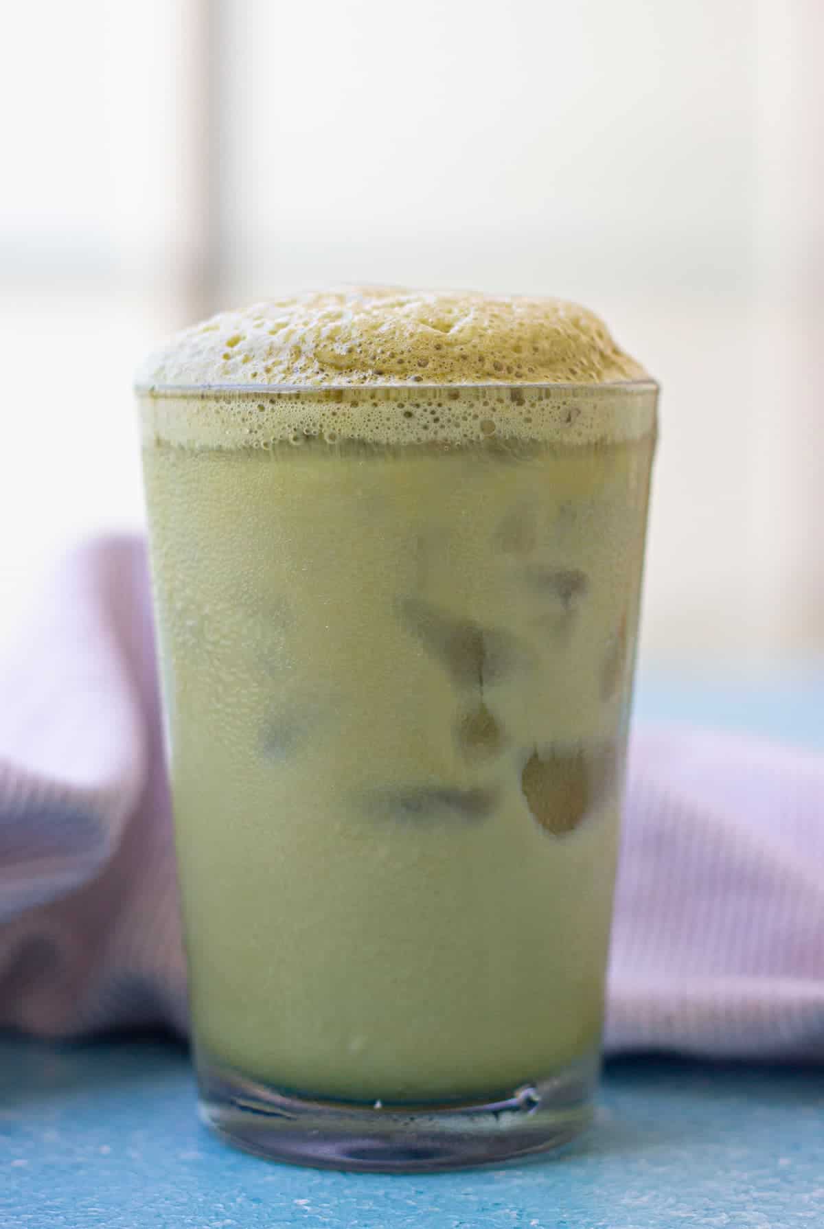 A glass of iced matcha latte topped with frothy foam sits on a blue surface with a light purple cloth in the background.