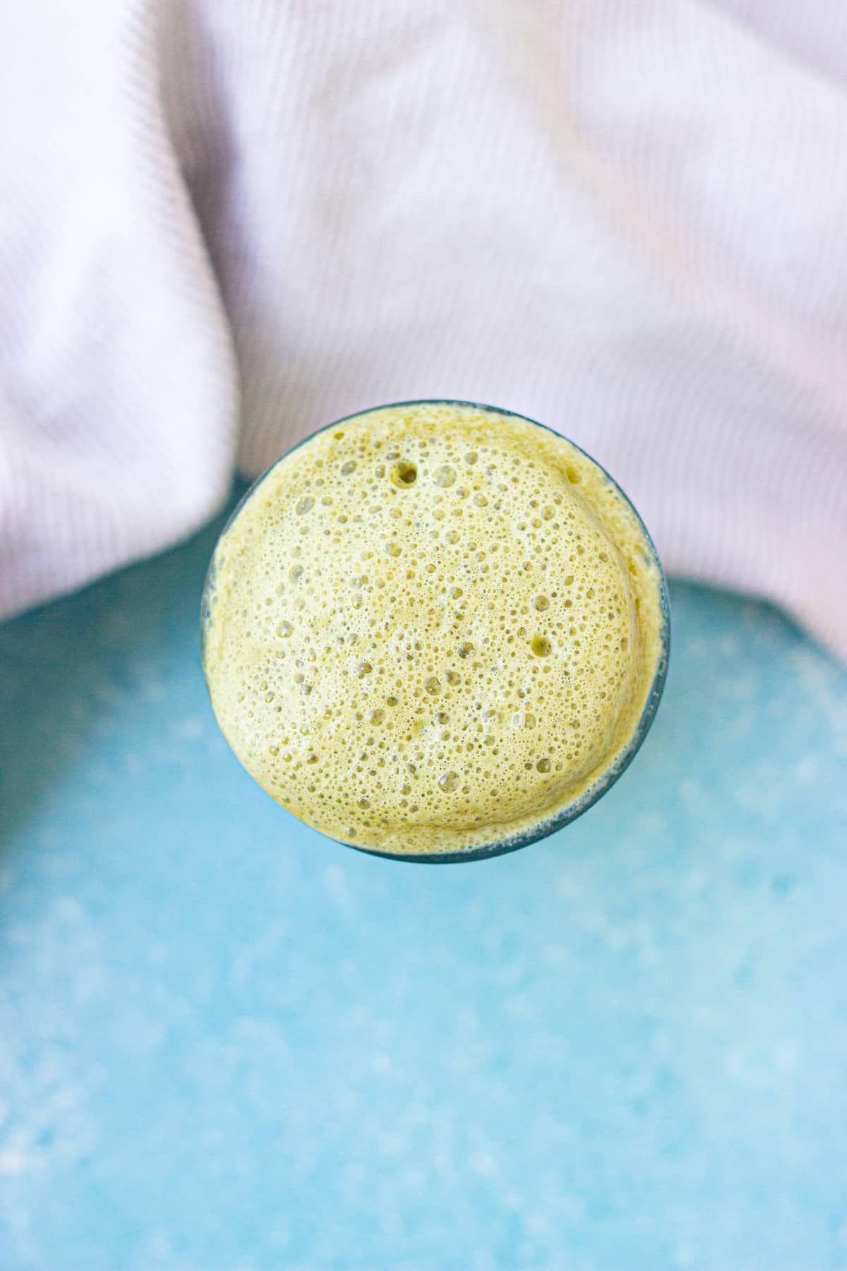 A glass filled with a frothy green beverage, viewed from above, set on a blue surface with a textured light cloth in the background.