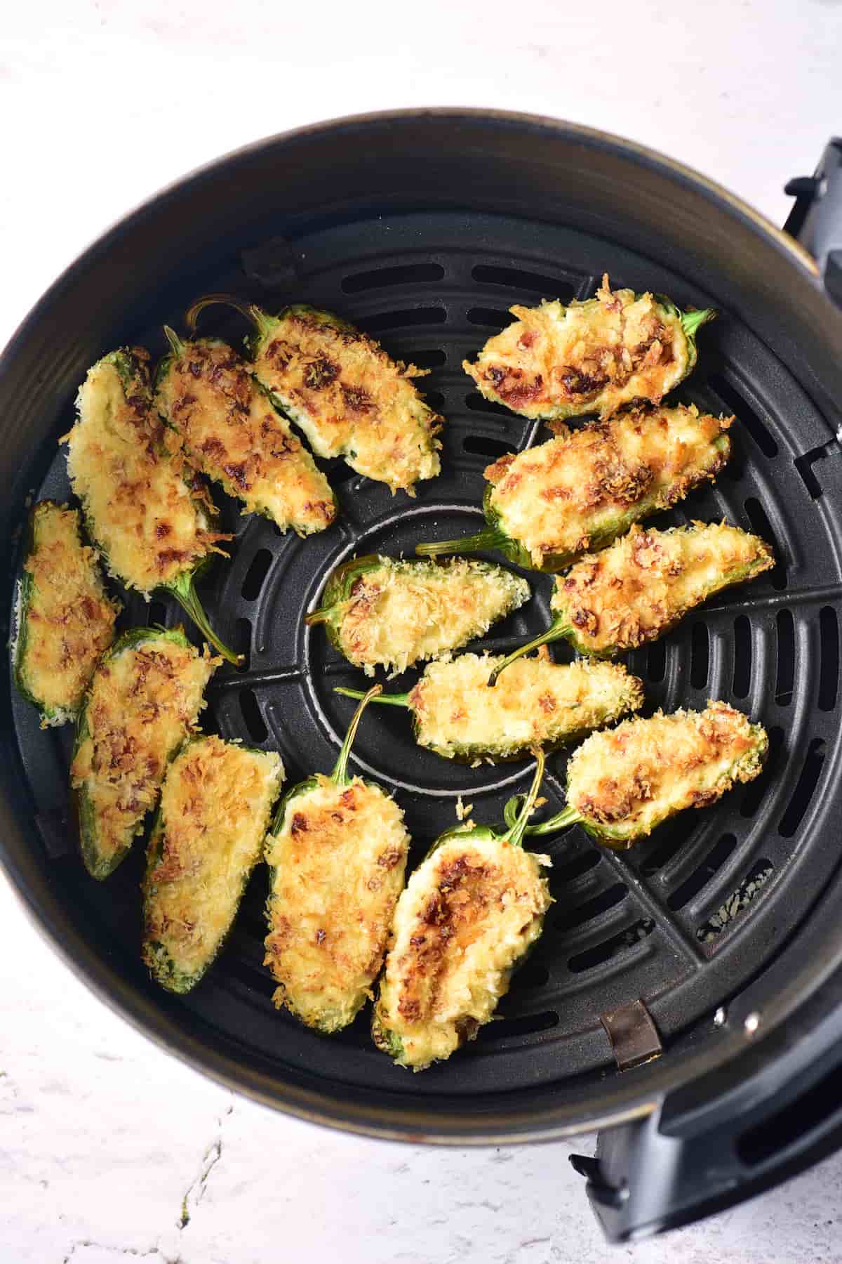 Halved jalapeños filled with a breadcrumb and cheese mixture are arranged in an air fryer basket, cooked until golden brown on top.