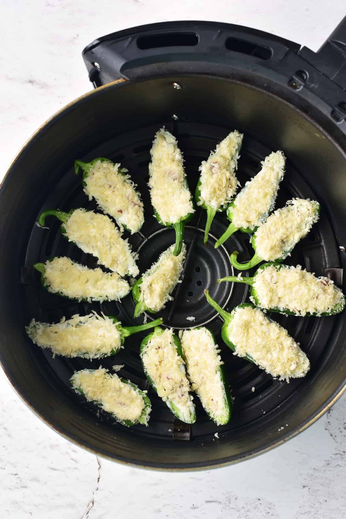Halved jalapeños stuffed and coated with breadcrumbs are arranged in a single layer inside an air fryer basket, ready to be cooked.