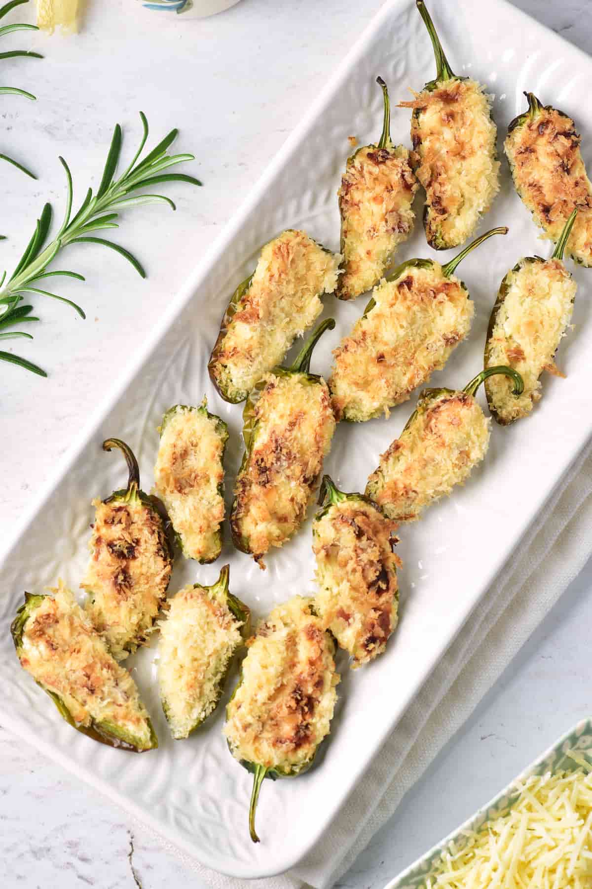 A white rectangular plate with baked, breadcrumb-topped jalapeño poppers arranged neatly on a white surface, garnished with a rosemary sprig.