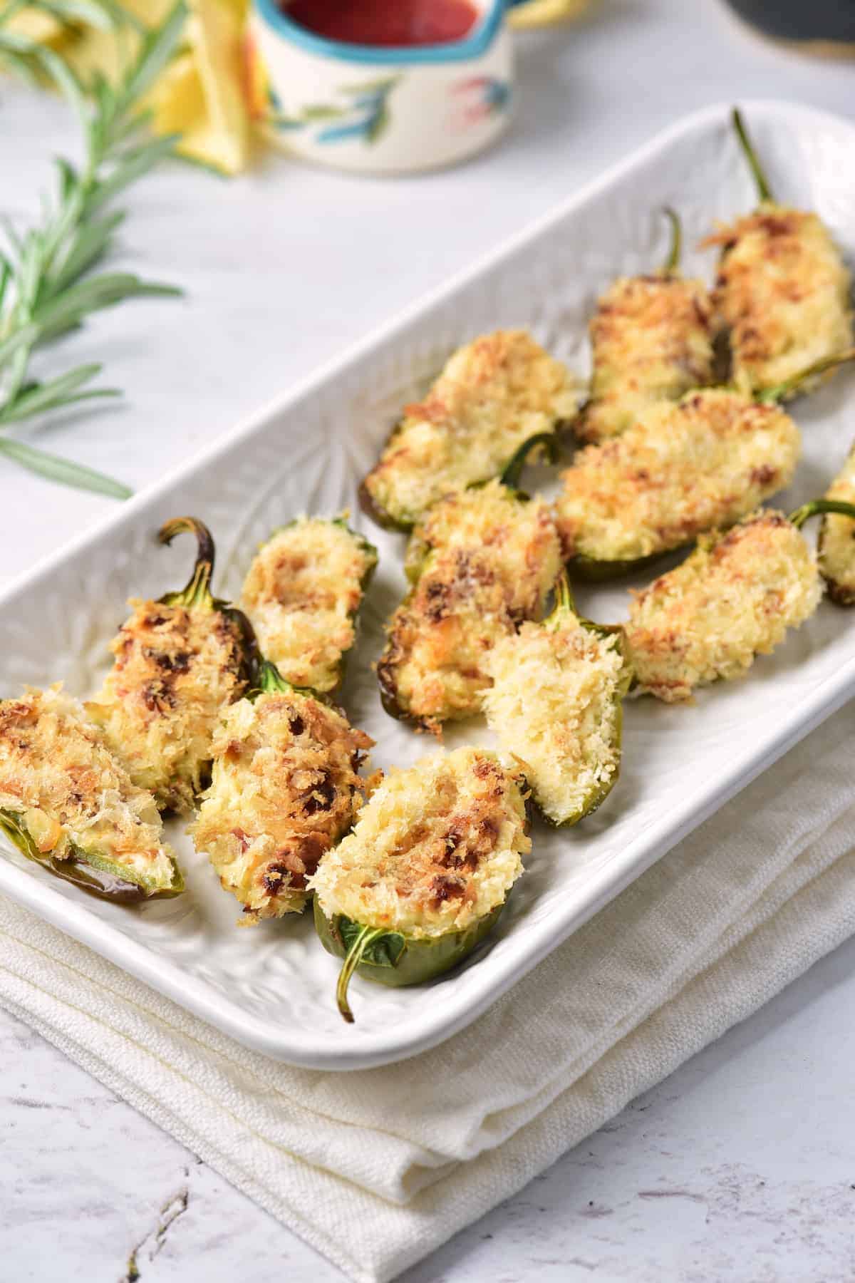 A rectangular white plate holds several baked, breadcrumb-topped stuffed jalapeño halves, arranged neatly on a light-colored surface with a napkin underneath.