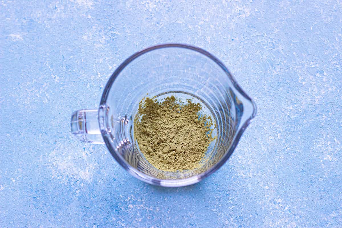Clear glass measuring cup containing a small amount of greenish powder, placed on a textured light blue surface.