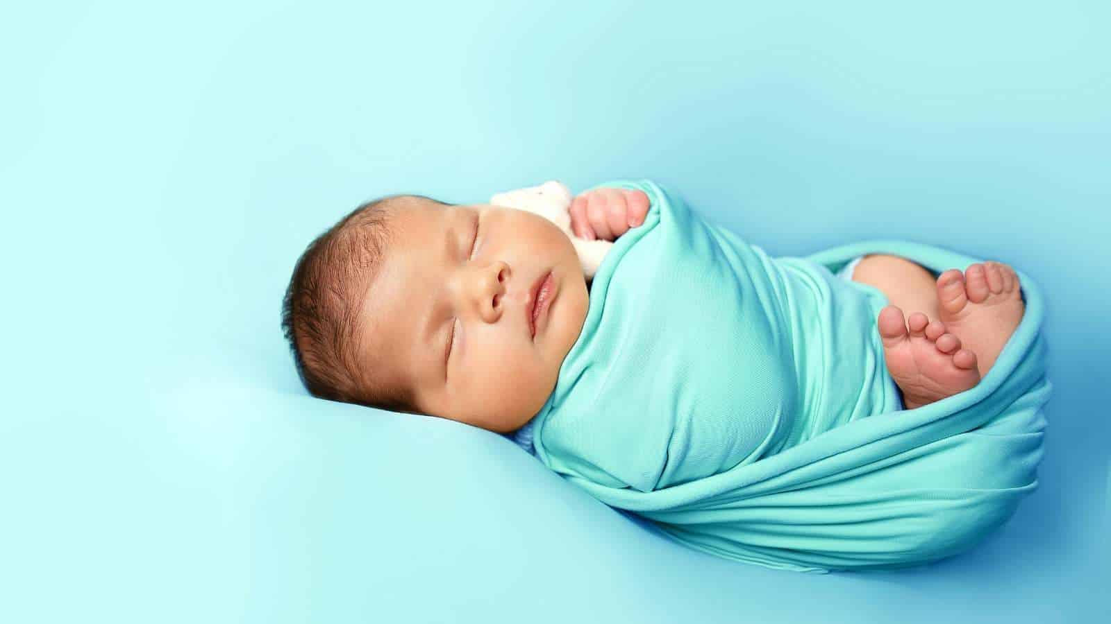 Newborn baby sleeping peacefully, wrapped in a light blue blanket against a matching blue background.