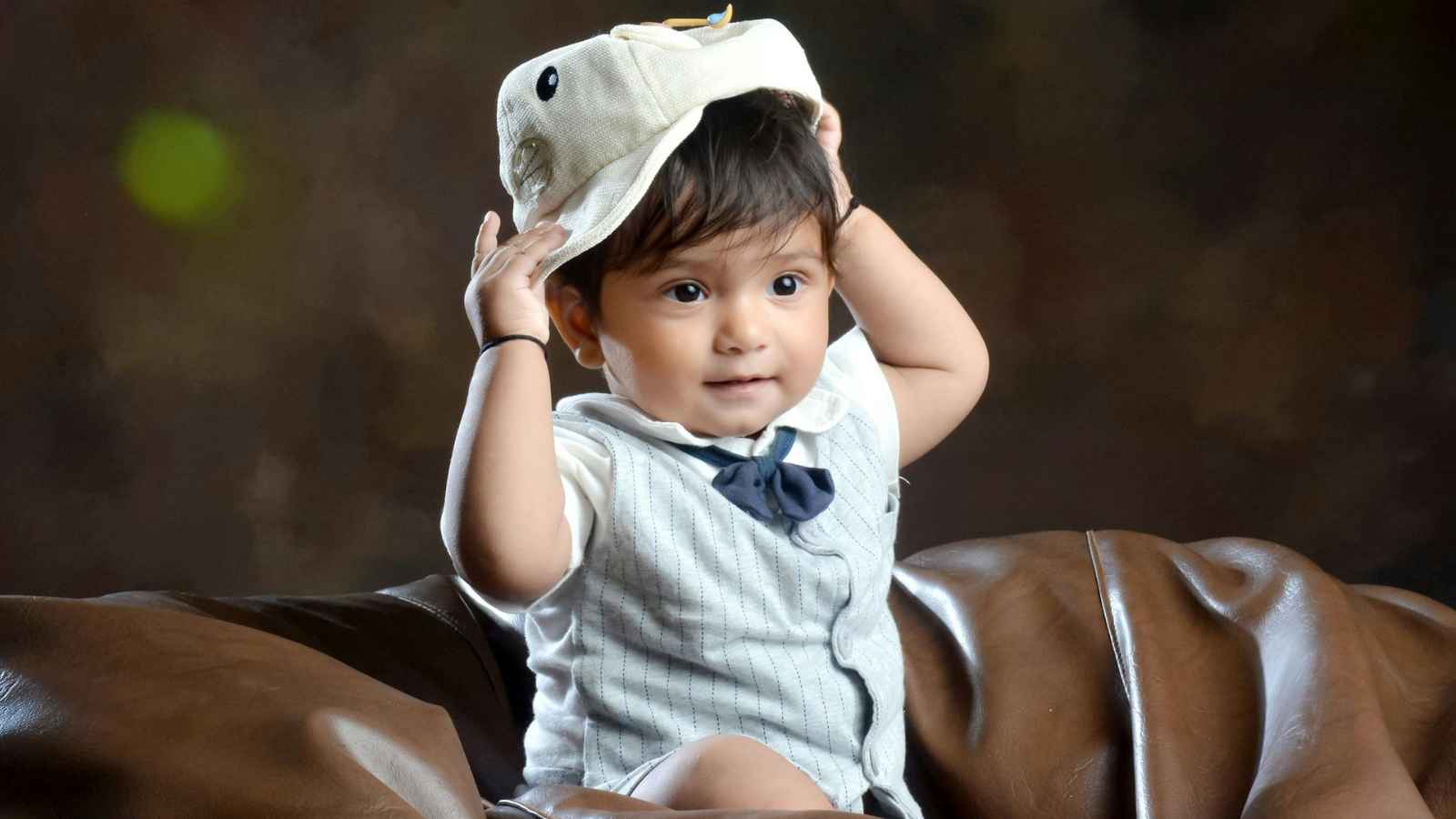 A young child in formal attire, sits on a brown leather chair, lifting a light-colored hat off their head and looking slightly to the side.