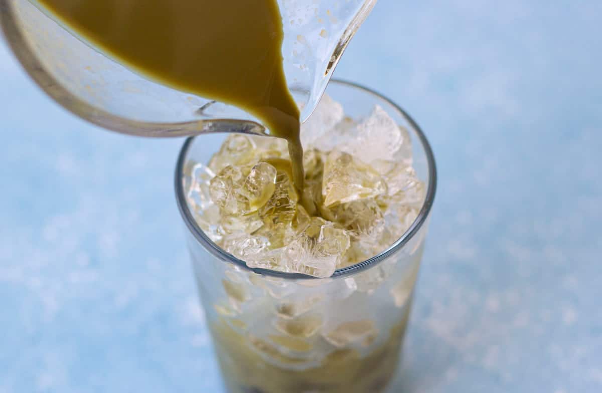 A glass filled with ice is being poured with a green liquid from a pitcher against a light blue background.