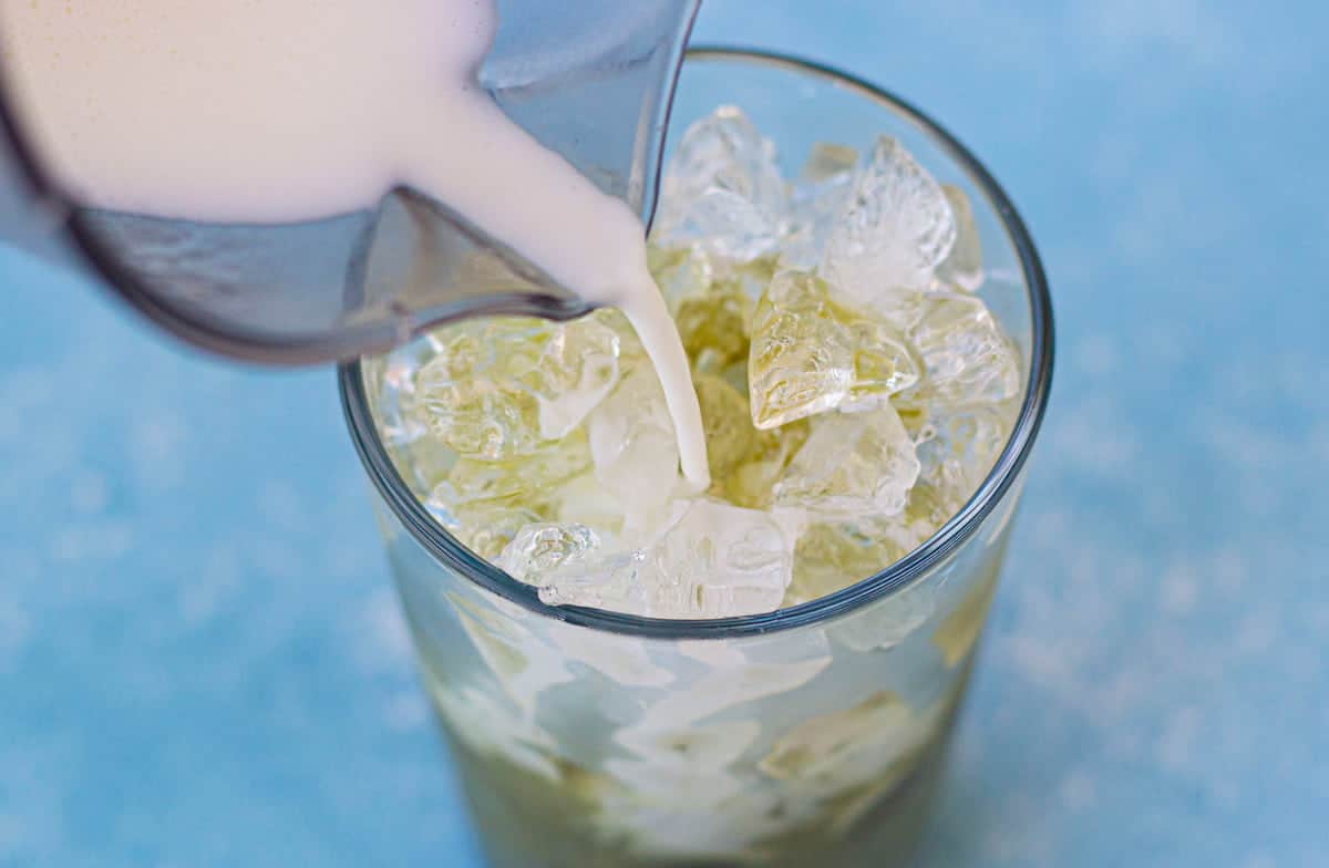 Milk is being poured from a pitcher into a glass filled with ice and a green liquid, against a blue background.