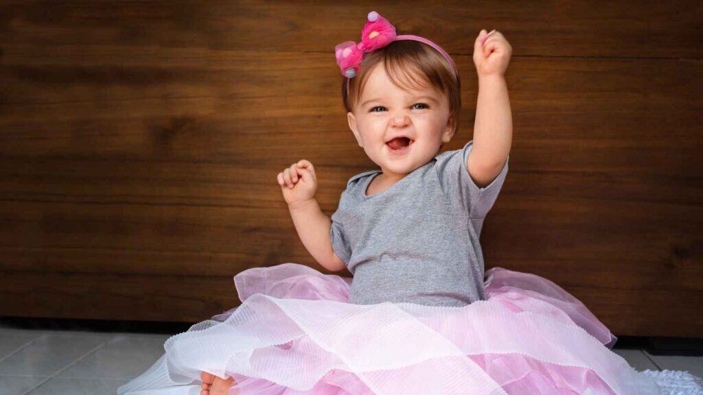 A baby wearing a grey shirt, pink headband, and fluffy pink skirt sits on the floor, smiling and raising one arm.