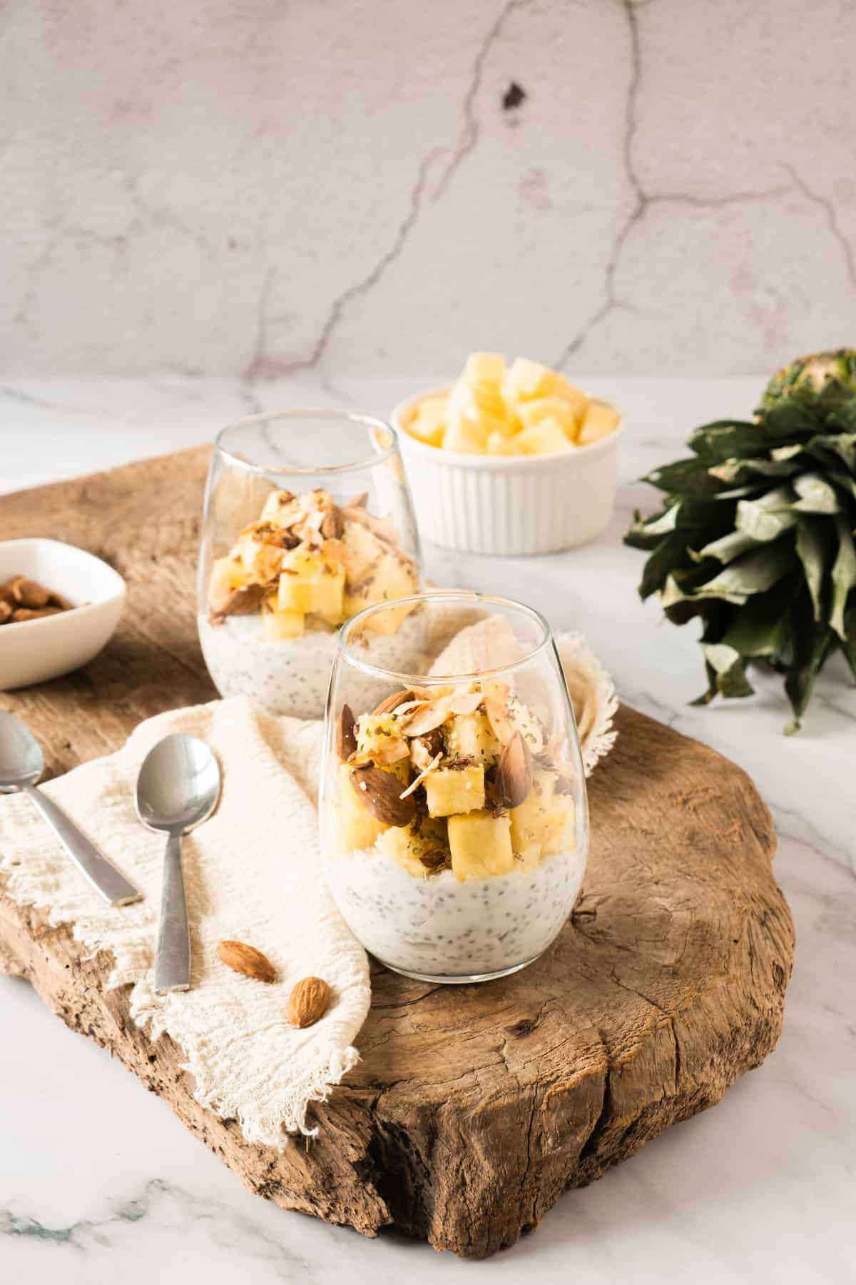 Two glasses of chia pudding topped with pineapple chunks and nuts on a wooden board, with a bowl of pineapple, almonds, a napkin, and spoons nearby.