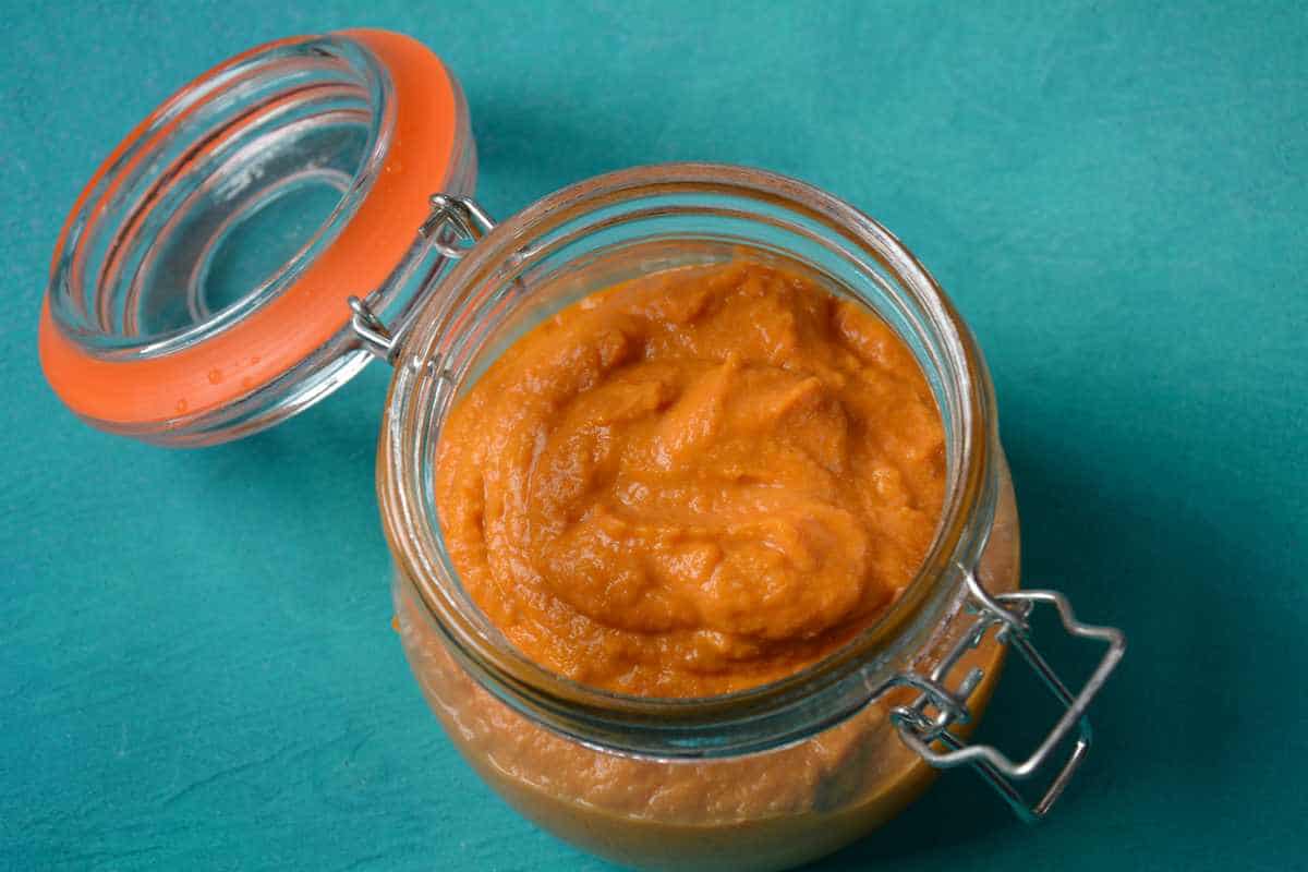 A glass jar with an orange puree, possibly pumpkin or carrot, sits open on a teal surface.