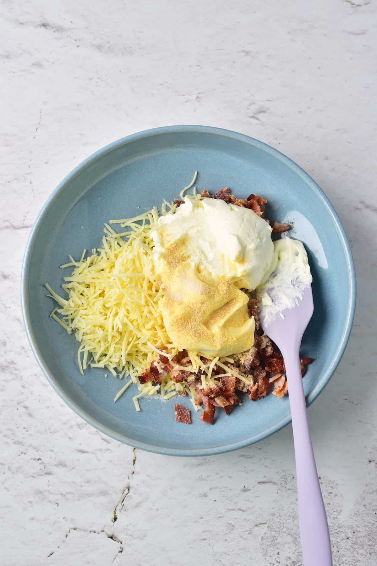 A blue bowl contains shredded cheese, bacon bits, a scoop of sour cream, and a sprinkle of seasoning, with a purple spatula resting inside.