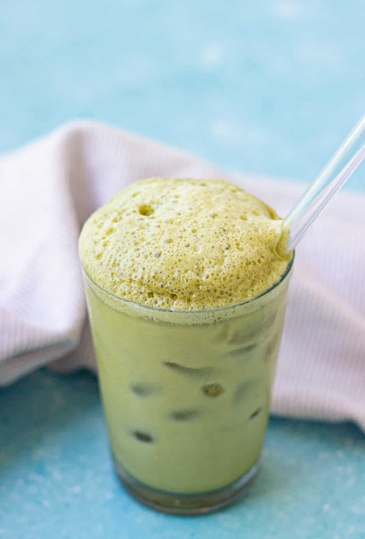 A glass of iced matcha latte with foam on top and a clear straw, placed on a blue surface with a striped cloth in the background.