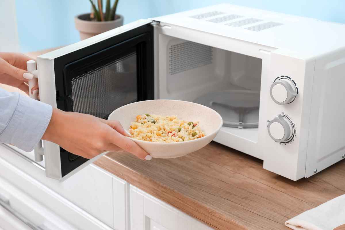 A person is placing a bowl of rice with vegetables into a white microwave on a kitchen counter.